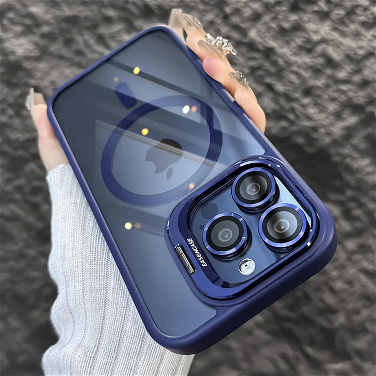 Close-up of a smartphone with a clear blue case, highlighting three camera lenses. The device is held by a hand wearing a light gray sweater with textured gray background.