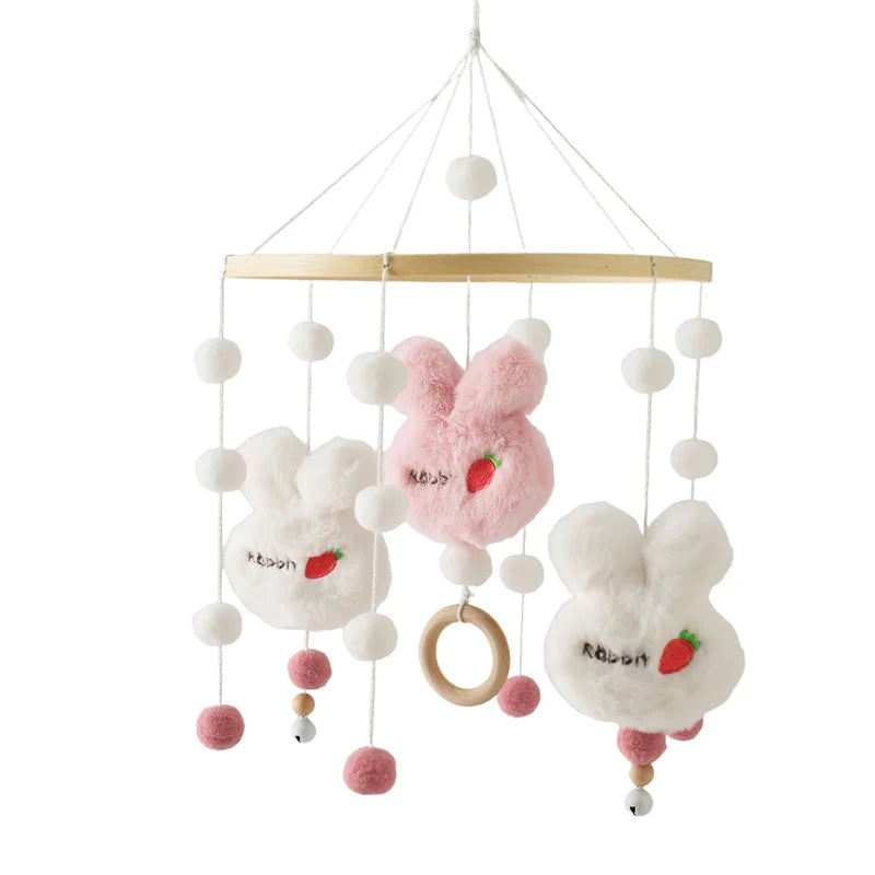 Baby mobile featuring plush bunny heads in white and pink with strawberry embroidery, hanging from strings with white and pink pom-poms, a wooden hoop, and small bells.
