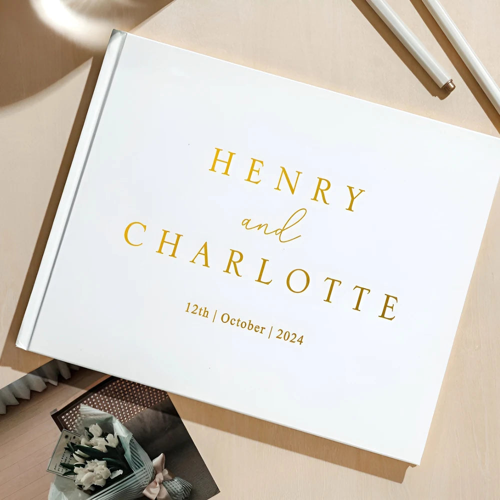 White photo album with "Henry and Charlotte" and "12th October 2024" in gold lettering. It lies on a wooden table next to a pen and a small bouquet.