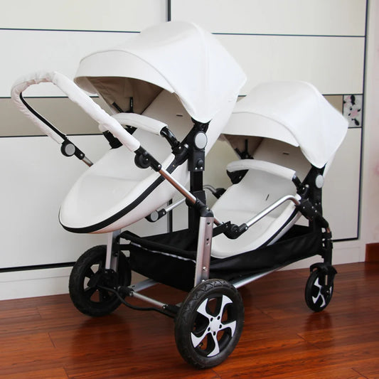 White double stroller with two seats side by side, featuring canopies and a padded handle. It has large wheels and a sturdy frame on a wooden floor background.