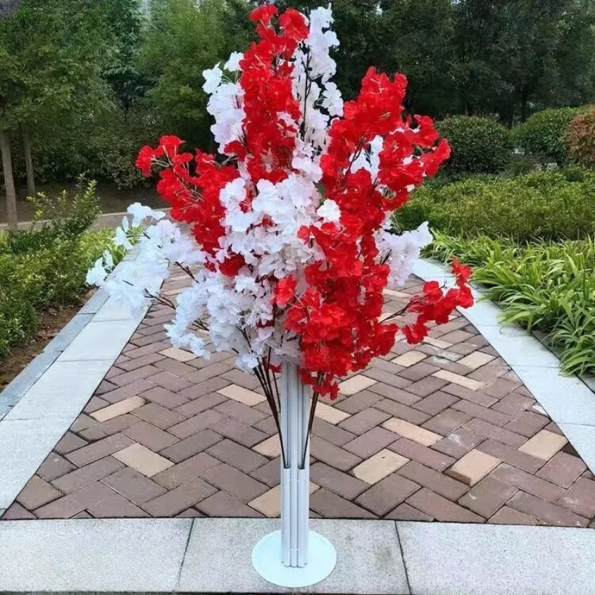 Artificial red and white flowers in a tall, white vase placed on a paved path. Green shrubs and trees surround the area, creating a vibrant and colorful outdoor display.