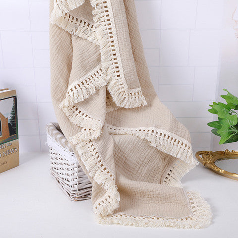 A beige textured blanket with fringe edges drapes over a white wicker basket on a white surface, next to a potted plant and partially visible book against a tiled wall.