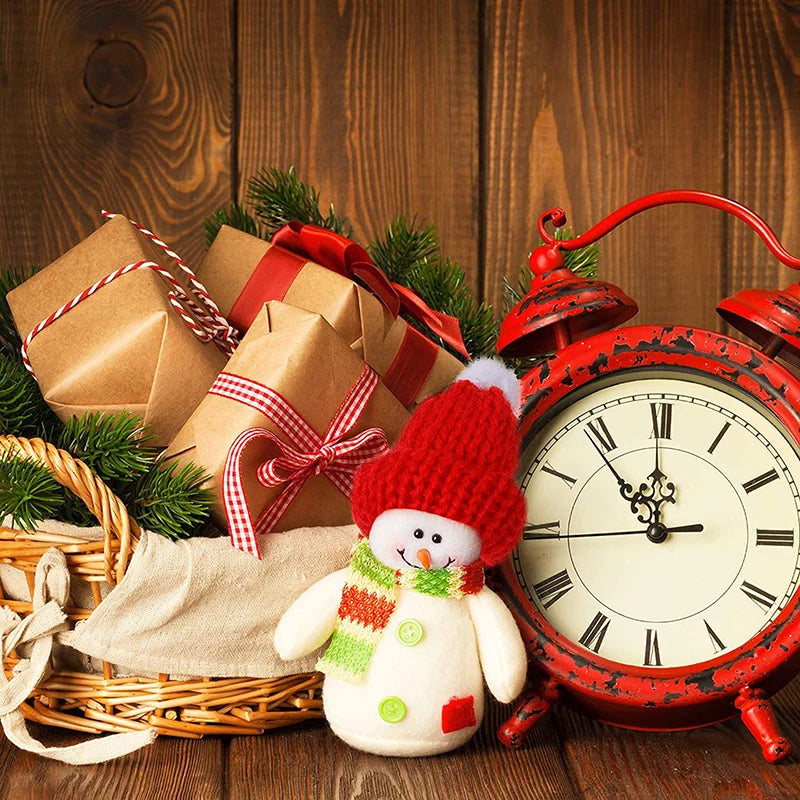 A vintage red alarm clock, wrapped gifts with red ribbons, and a smiling snowman doll in a red hat and green scarf sit in a basket with evergreen branches on a wooden background.