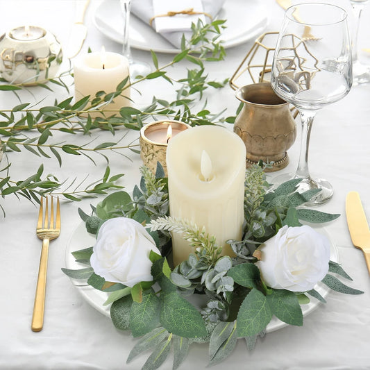 Elegant table setting with a white candle surrounded by white roses and greenery. Gold cutlery and a wine glass complement the decor on a white tablecloth.