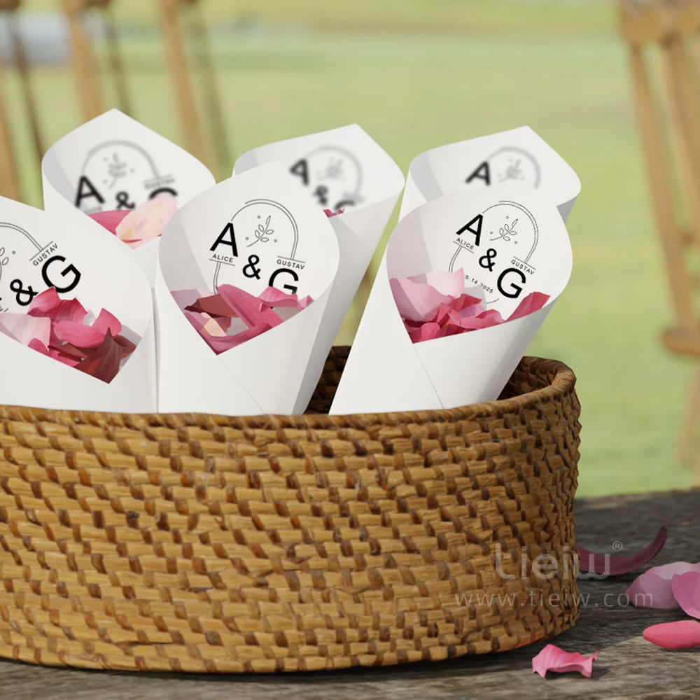 A woven basket holds white paper cones filled with pink petals. Each cone is labeled "A & G" with a floral design, set on an outdoor wooden table.