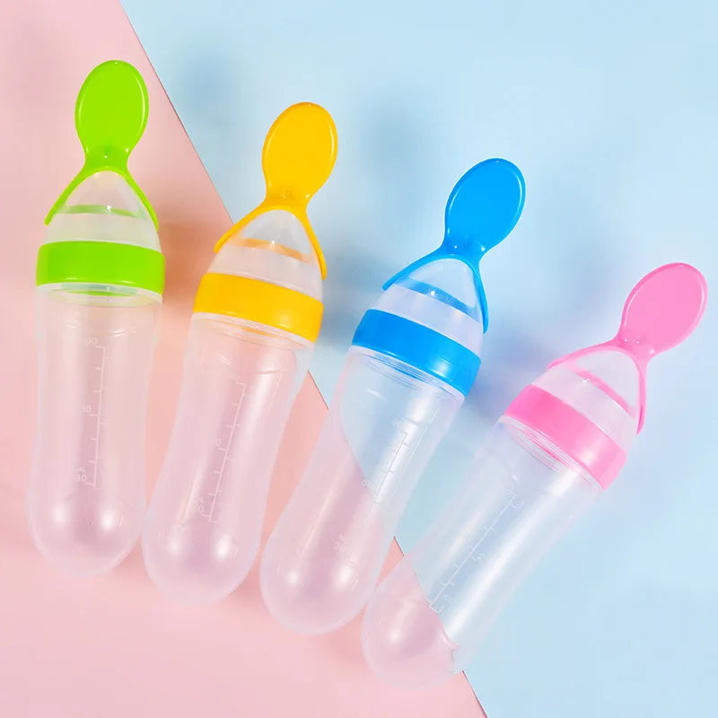 Four baby feeding bottles with attached spoons, each with a different color cap: green, yellow, blue, and pink. The background is split diagonally into pink and blue.