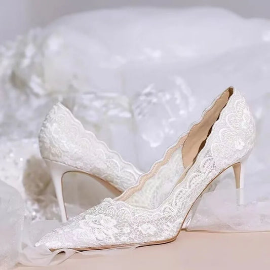 Elegant white lace heels with pointed toes and scalloped edges, set on a soft, white fabric background.