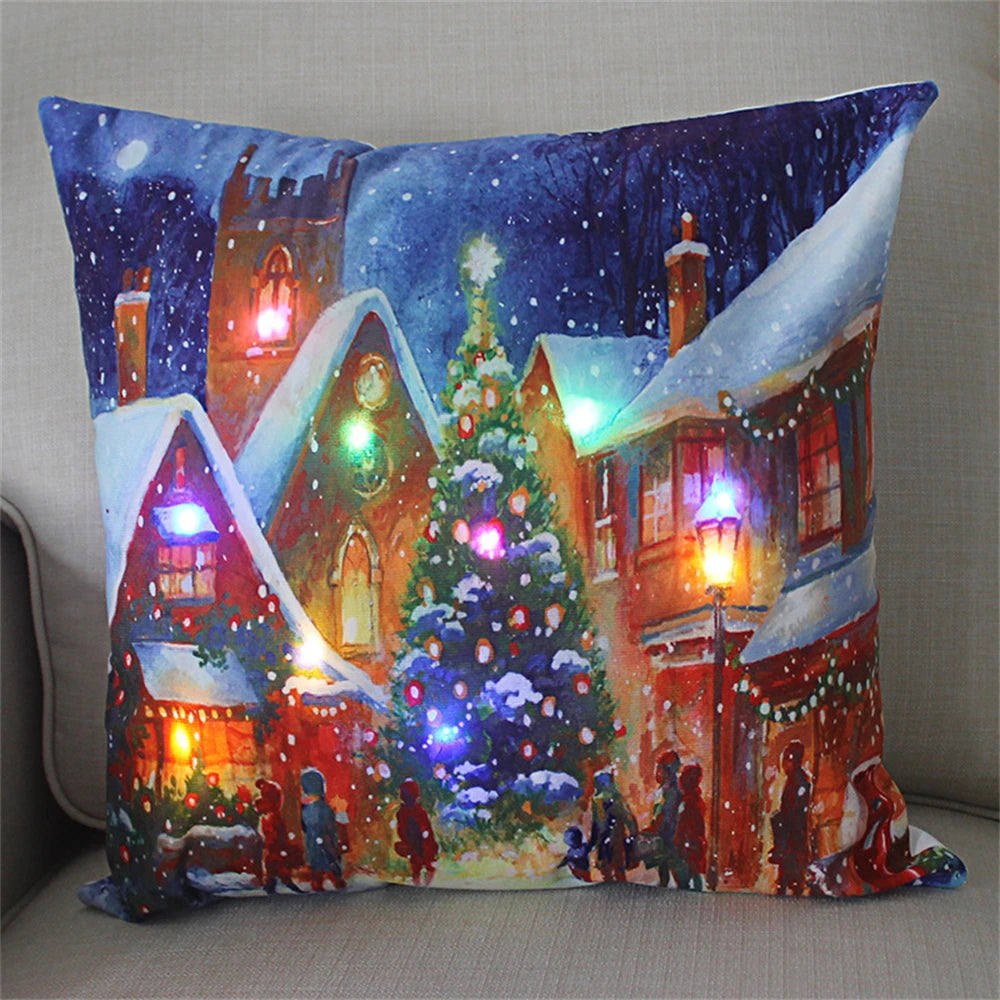 Festive pillow depicting a snowy village scene with lit houses and a decorated Christmas tree. Warm, multicolored lights create a cozy, holiday atmosphere.