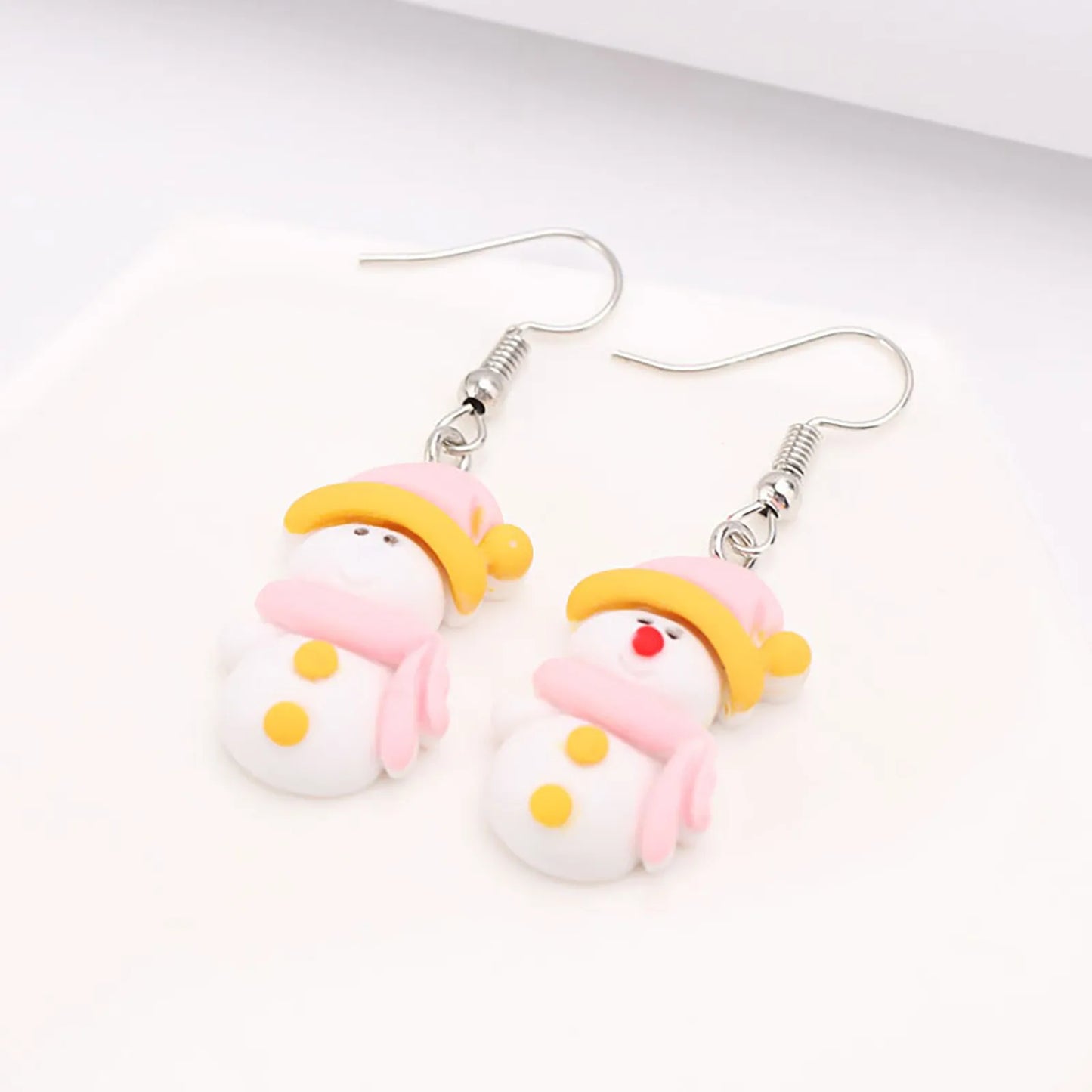 Cute snowman earrings with pink hats and scarves, yellow noses and buttons, and silver hooks, set on a light background.