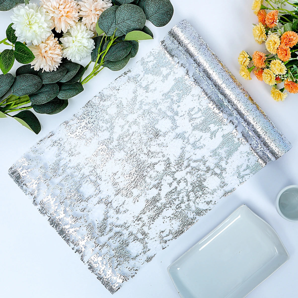 Silver and white textured table runner on a white surface, flanked by green leaves with cream and orange flowers, alongside a grey plate and cup.