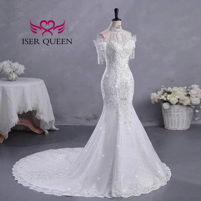 Elegant white fishtail bridal gown on a mannequin, adorned with lace and beadwork, featuring shoulder fringes. Background shows a table with a floral arrangement and a gray wall.