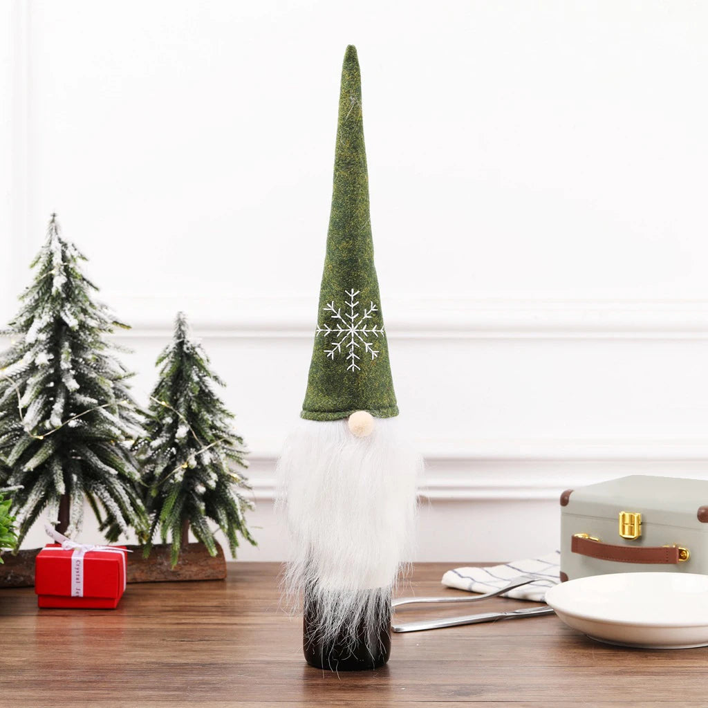 Festive gnome bottle topper with green hat and snowflake design, white beard, on a wooden table. Small snowy trees and a wrapped gift in the background add a holiday touch.