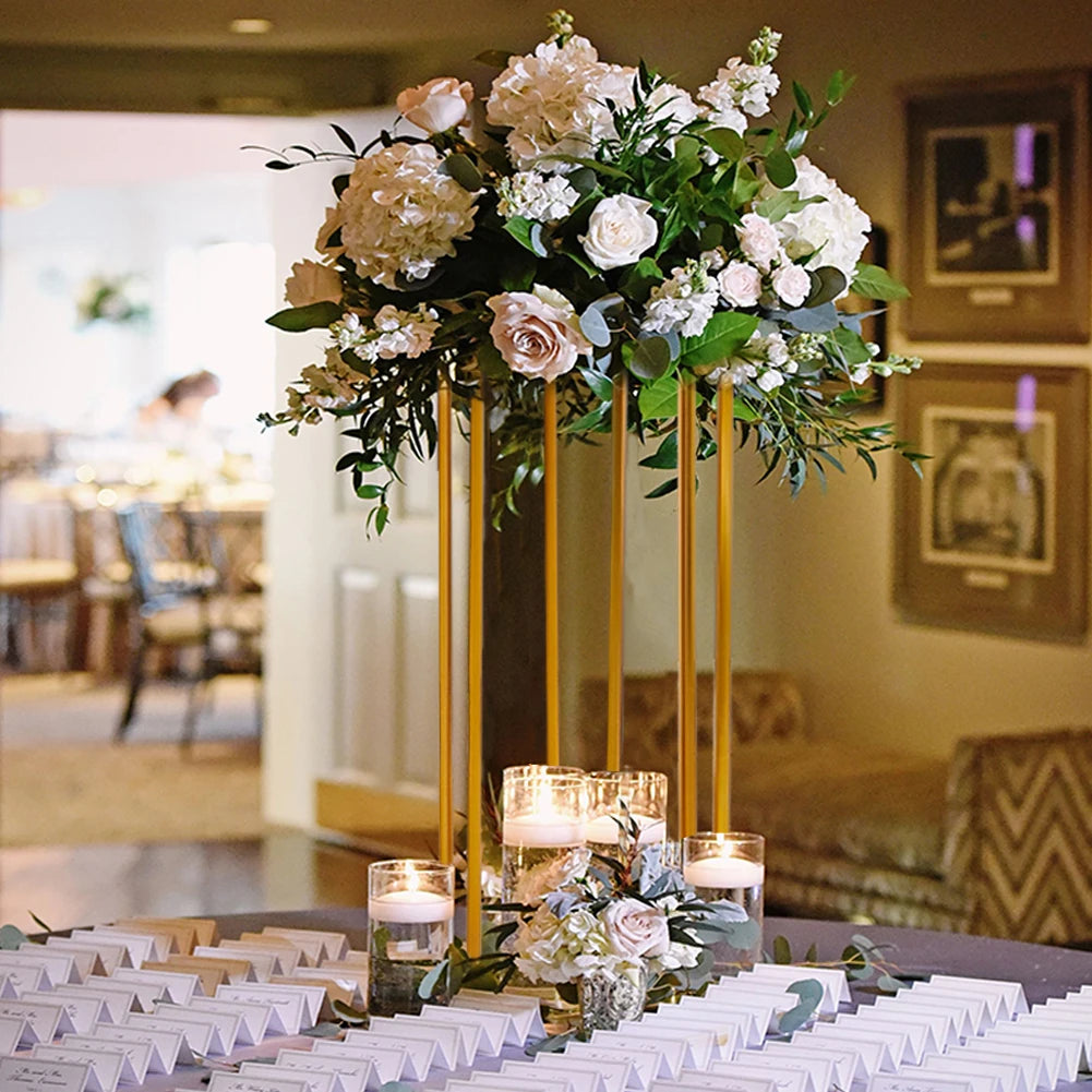 Elegant flower arrangement on a tall stand with white roses and greenery, surrounded by candles on a table with place cards. Cozy, warmly lit room in the background.