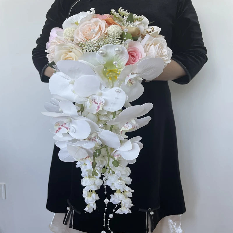 A person in a black dress holds a cascading bouquet with white orchids, pink and peach roses, and delicate greenery. Pearls accent the arrangement, adding elegance.