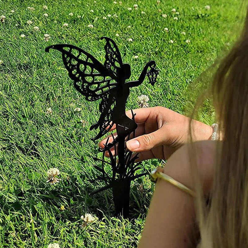 A child's hand places a decorative metal fairy silhouette with detailed wings into lush green grass dotted with white clover flowers, basking in sunlight.