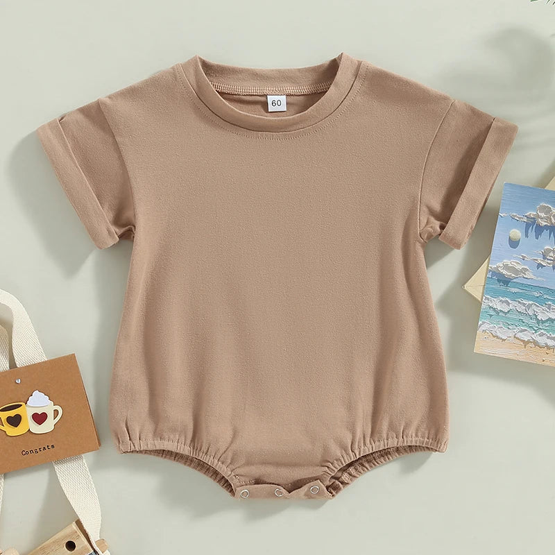 A light brown baby romper with short sleeves, featuring a round neck and snap closures at the bottom. Surrounding it are a card with two mugs and a seaside painting.