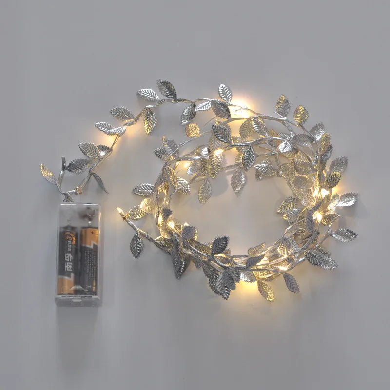 Silver leaf string lights glowing warmly, arranged in a coil. The attached battery pack is visible. The lights and leaves are designed to mimic a vine.