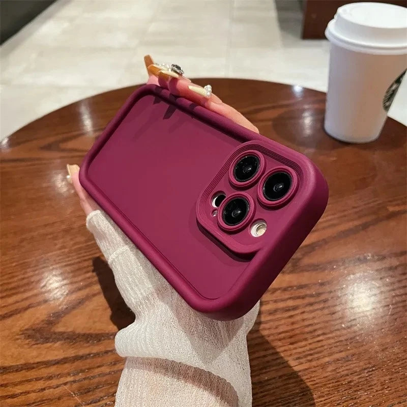 A hand with manicured nails holds a smartphone with a maroon case, featuring a prominent camera setup, against a wooden table with a cup in the background.