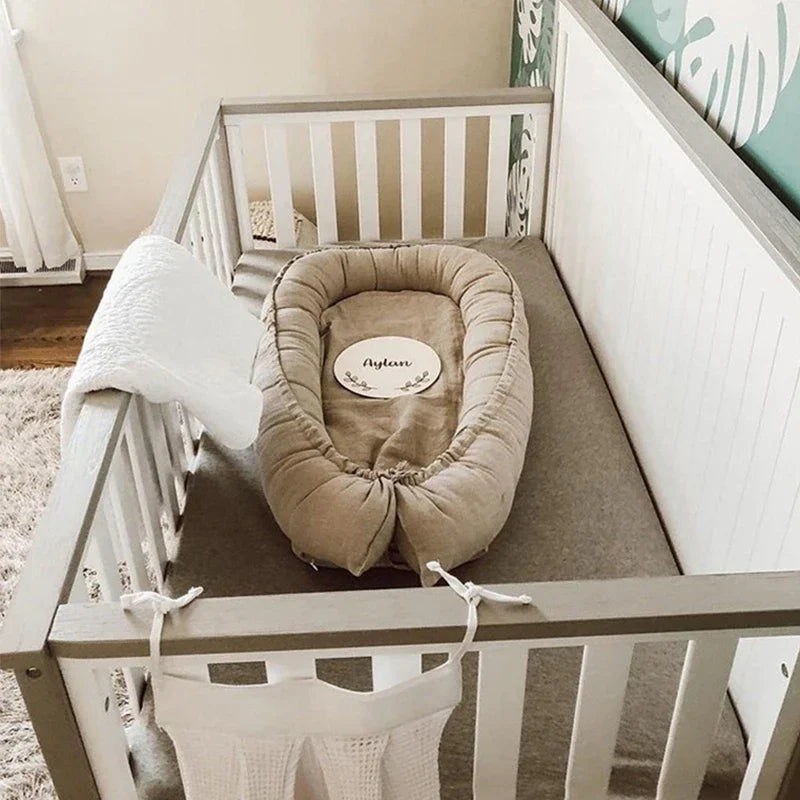 A cozy baby crib with a beige baby nest and nameplate, “Aylan,” inside. A white blanket hangs over the side. The nursery has soft lighting and leafy wallpaper.