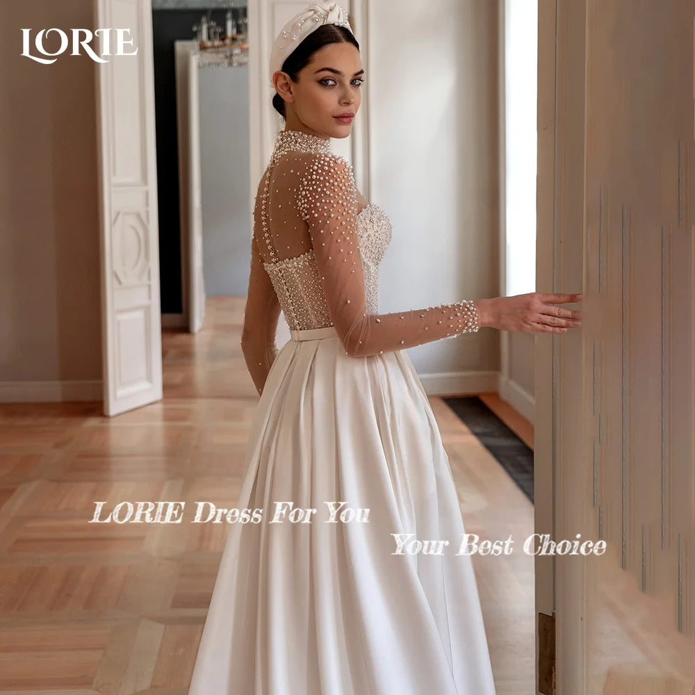 Elegant bride in a white gown with a pearl-embellished sheer top and long sleeves, standing in a softly lit room, looking back over her shoulder.