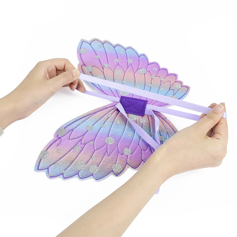 Hands holding a pair of colorful fairy wings in pastel shades of purple and blue with silver glitter dots, secured by light purple elastic straps.