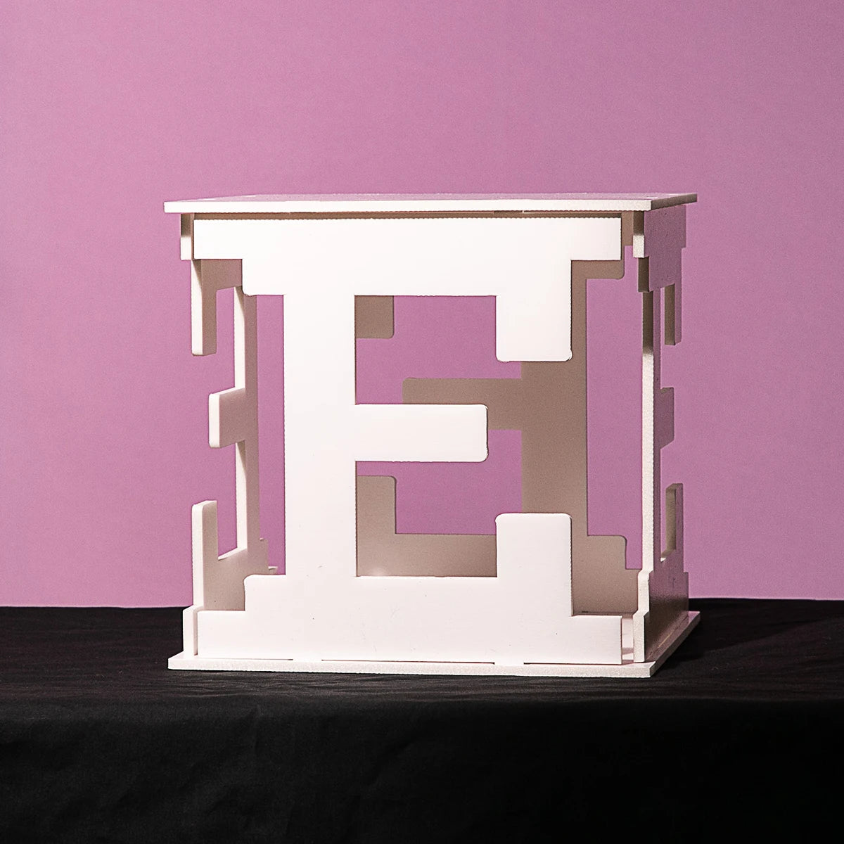 White 3D letter "E" sculpture with a geometric design on a black cloth surface. The background is a solid pink color.