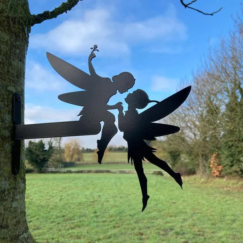 Silhouette of two fairy figures kissing atop a metal bracket mounted on a tree, set against a clear blue sky and a green, open field with distant trees.
