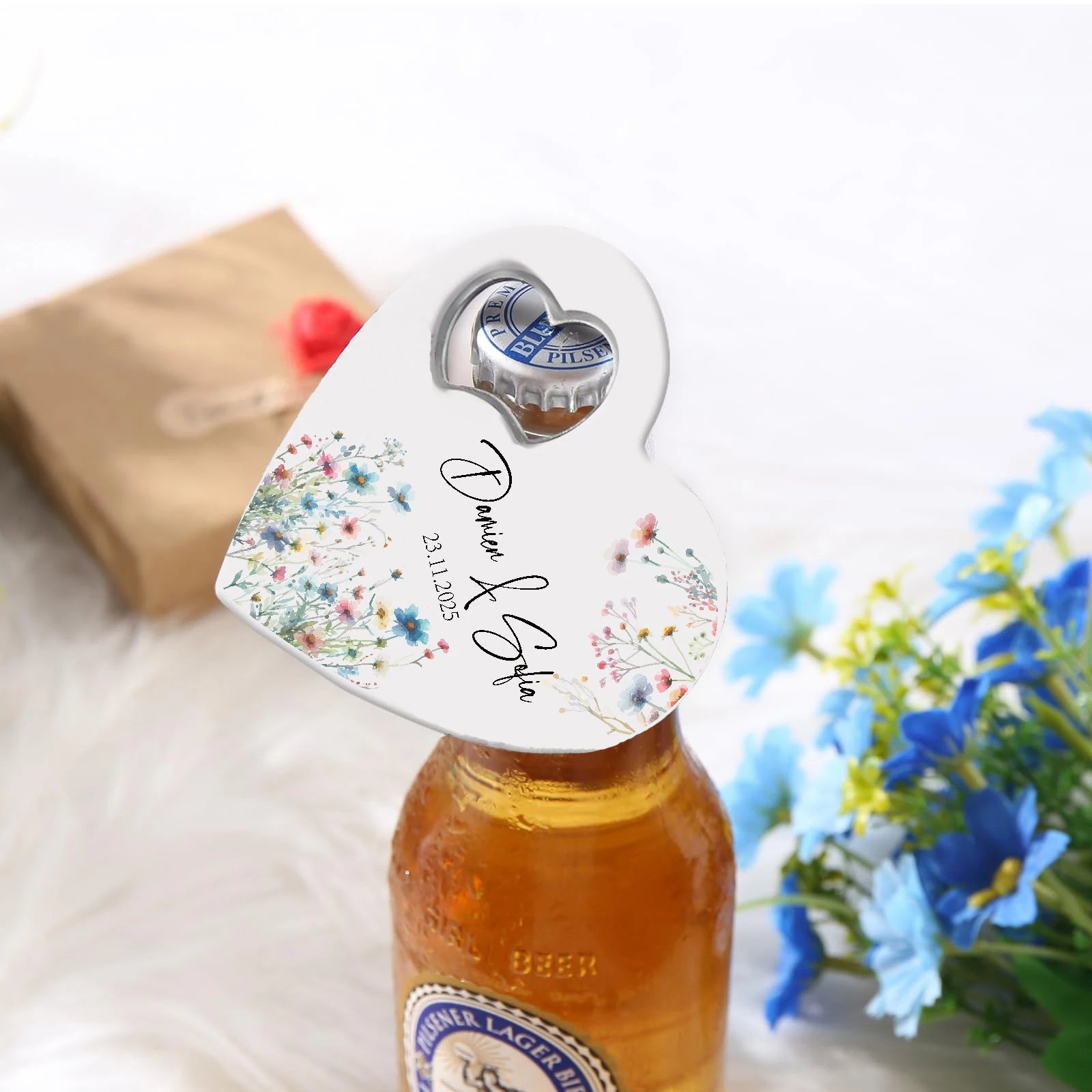 Heart-shaped bottle opener with floral design and names "Damien & Sofia" on a beer bottle. Background features a brown gift box and blue flowers on a white surface.