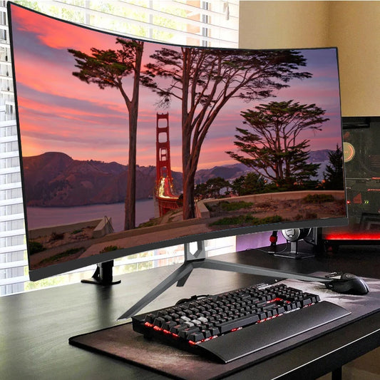 Curved monitor on a desk displays a sunset scene with trees framing a red bridge. Below, a black and red backlit keyboard sits on a large mouse pad beside a gaming mouse.