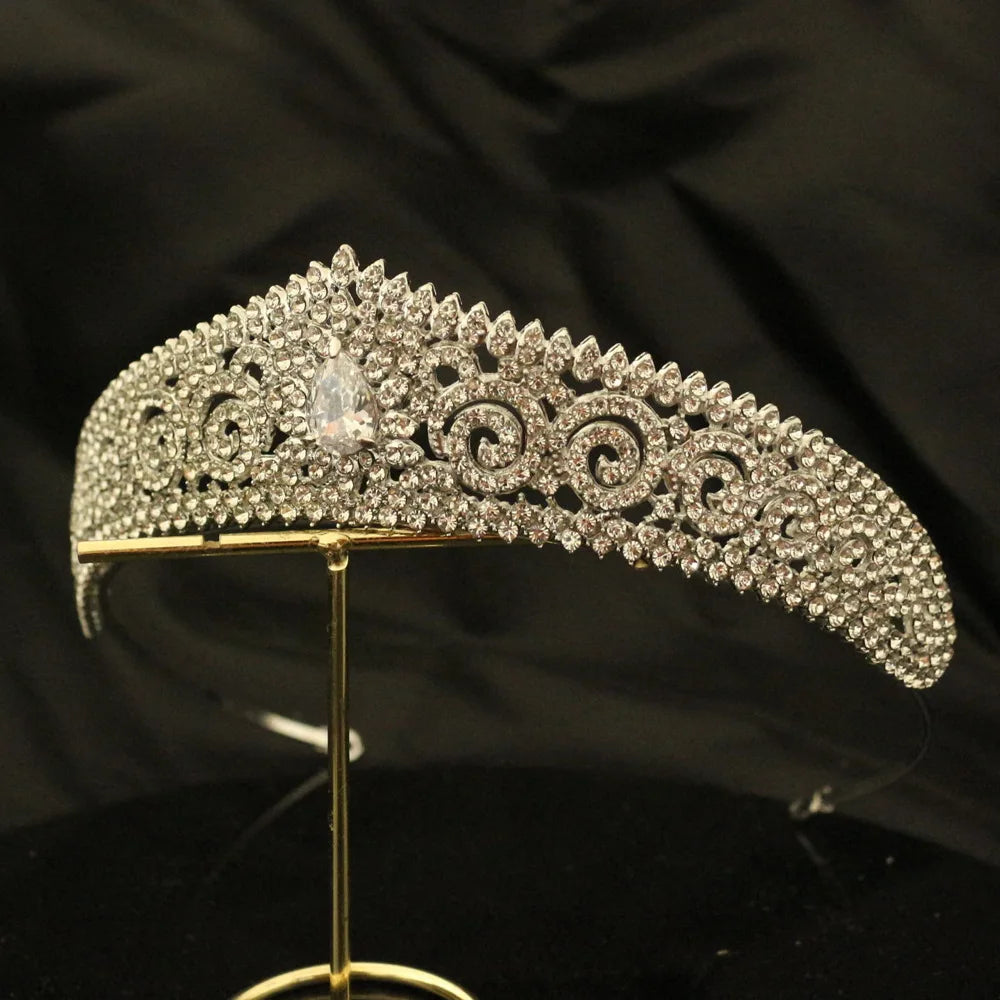 A sparkling tiara encrusted with intricate patterns of diamonds, featuring a prominent teardrop-shaped gem in the center, displayed on a gold stand against a dark background.