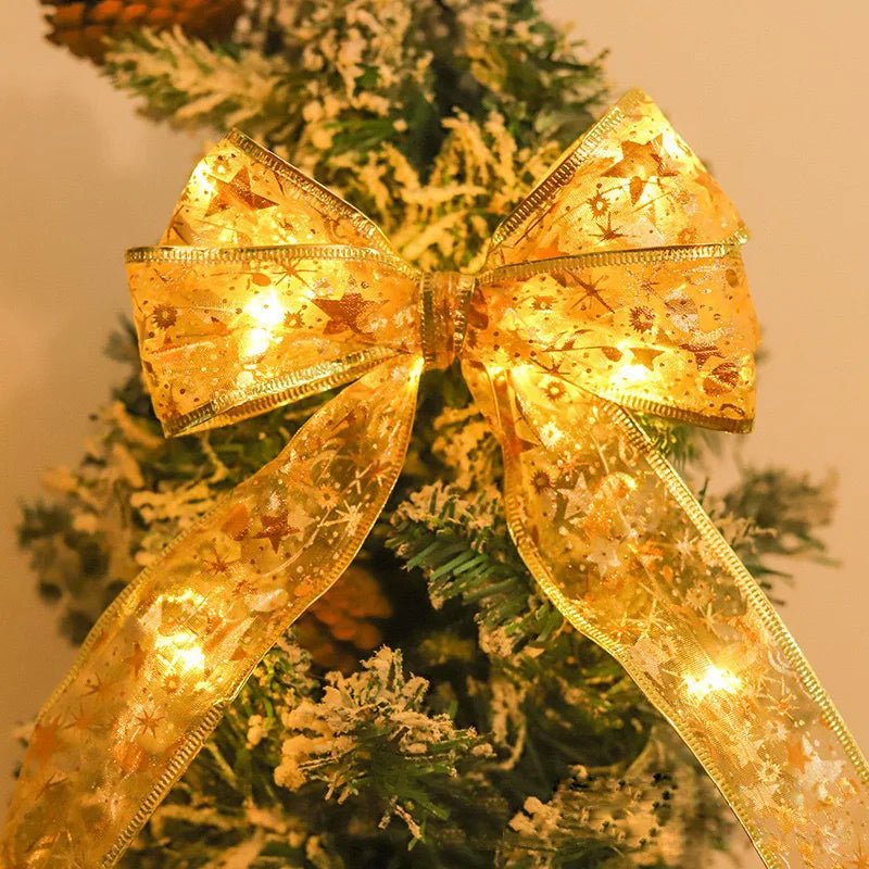 A Christmas tree adorned with a large, illuminated golden ribbon bow featuring stars and snowflakes, with snow-dusted branches in the background.