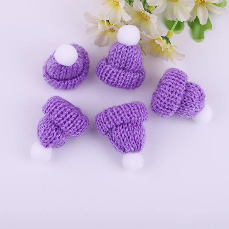 Five small, purple knitted hats with white pom-poms lie on a white surface. Yellow and white flowers are in the background, adding a touch of color.