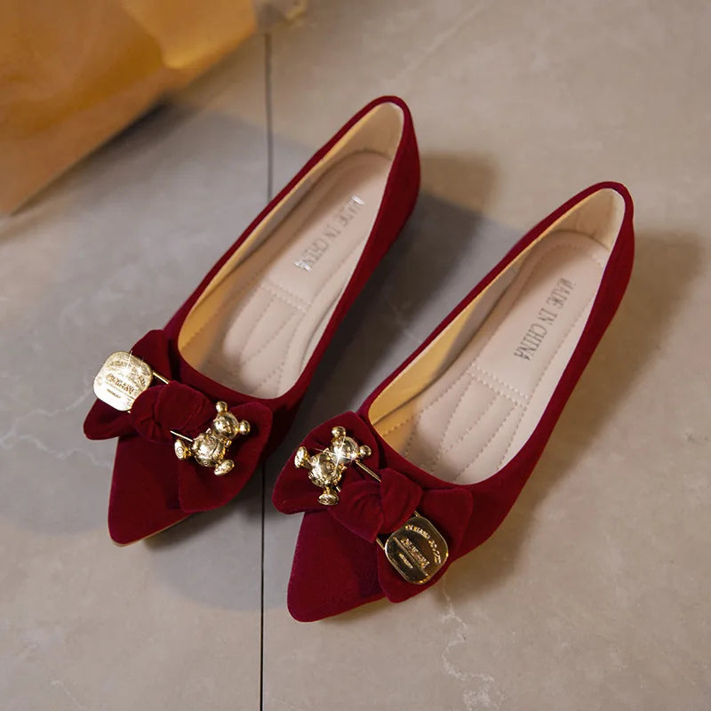 Red velvet flats with pointed toes, featuring gold bear and medallion embellishments on bows. The interior is cushioned, and the shoes rest on a tiled floor.