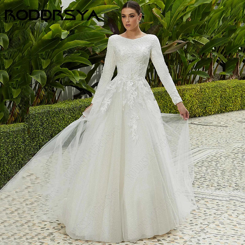 Woman in a long-sleeve, lace wedding dress with a flowing train stands on a patterned stone path, surrounded by lush green plants and neatly trimmed hedges.