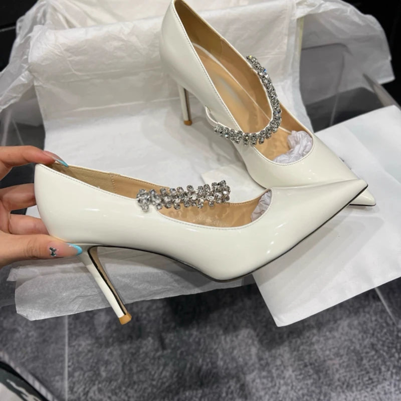 Elegant white high-heeled shoes with pointed toes and embellished with silver crystal straps. Shoes are displayed on tissue paper, with a hand holding one shoe.