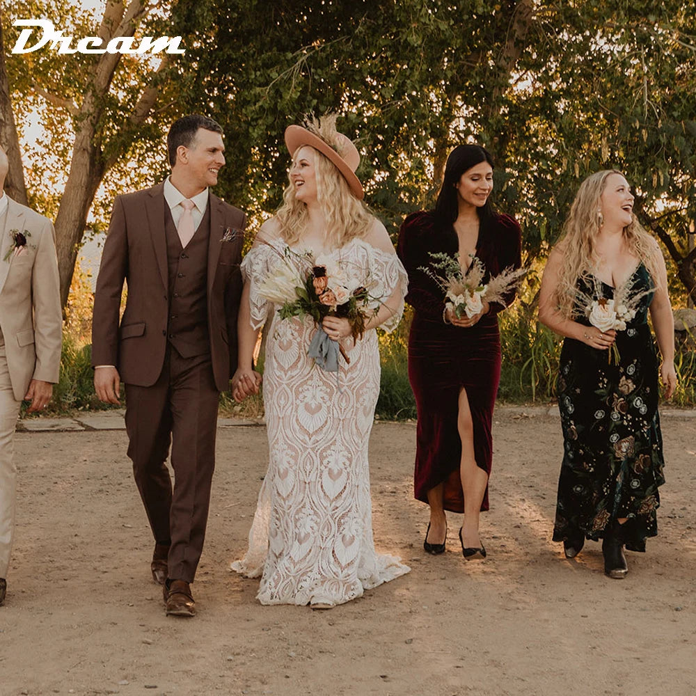 A bride in a lace gown and hat walks hand-in-hand with the groom in a brown suit. Bridesmaids in elegant dresses walk alongside, holding bouquets, with trees in the background.