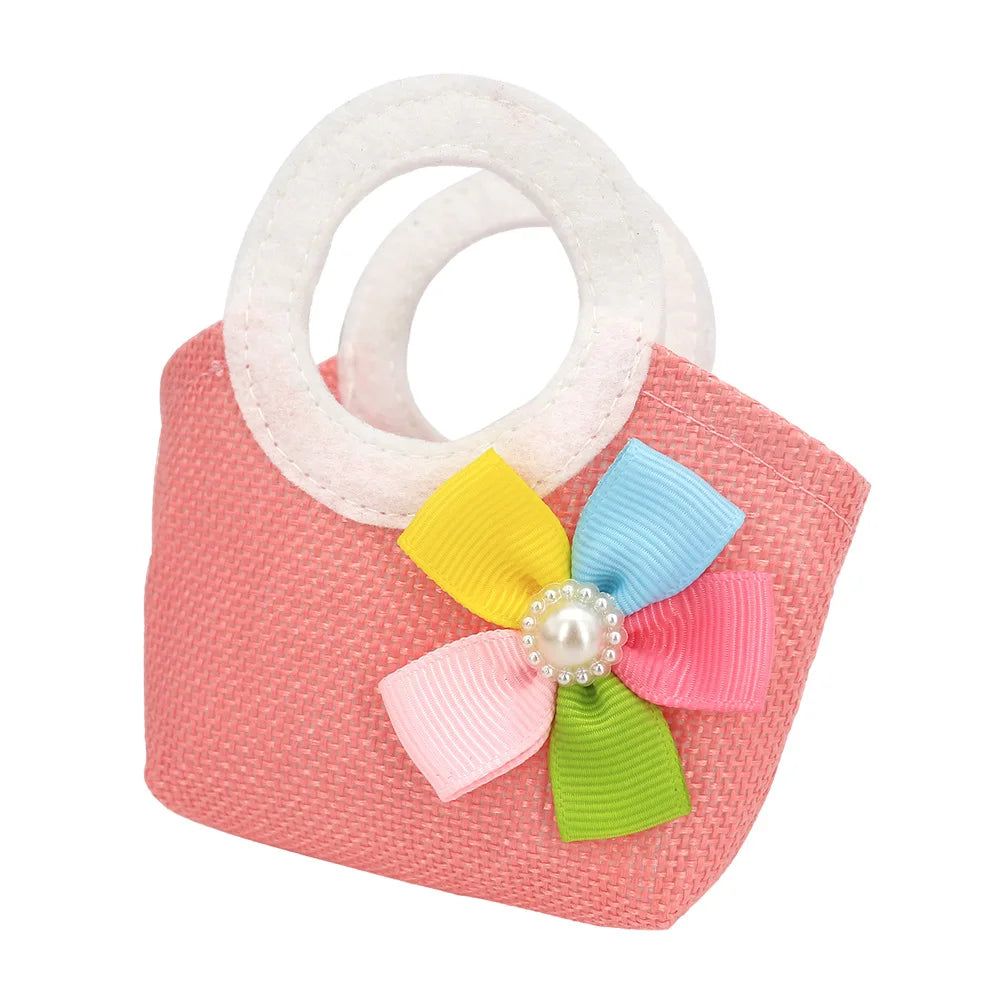 Small pink woven handbag with round handles, adorned with a colorful ribbon flower featuring petals in yellow, blue, pink, and green, centered by a faux pearl.