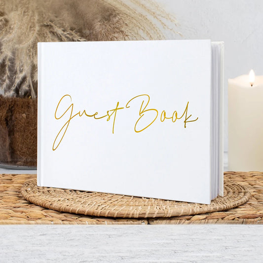 White guest book with "Guest Book" in gold script on a woven mat. Candle and pampas grass vase in the background add a cozy ambiance.