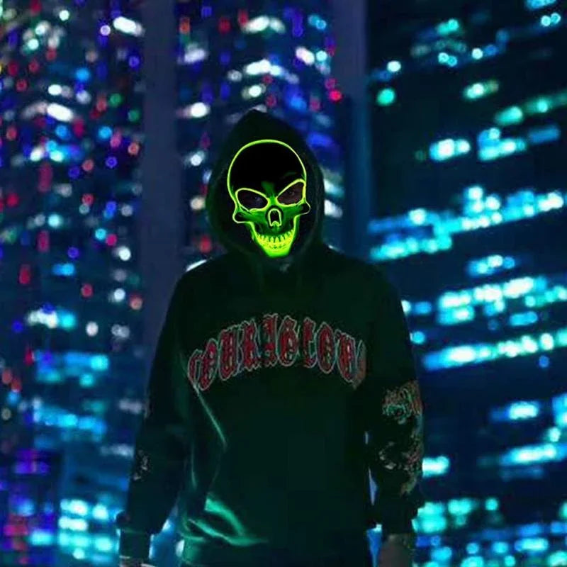 A person wearing a hoodie with a glowing green skull mask stands against a backdrop of colorful city lights at night.