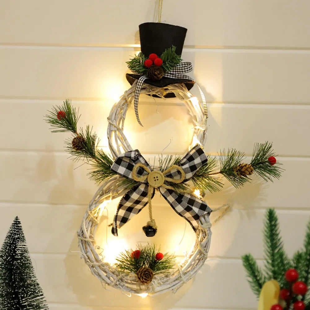 A lit snowman wreath hangs on a wall, featuring a black top hat, pine branches, red berries, and a black-and-white checkered bow with a wooden button. Festive decor surrounds it.