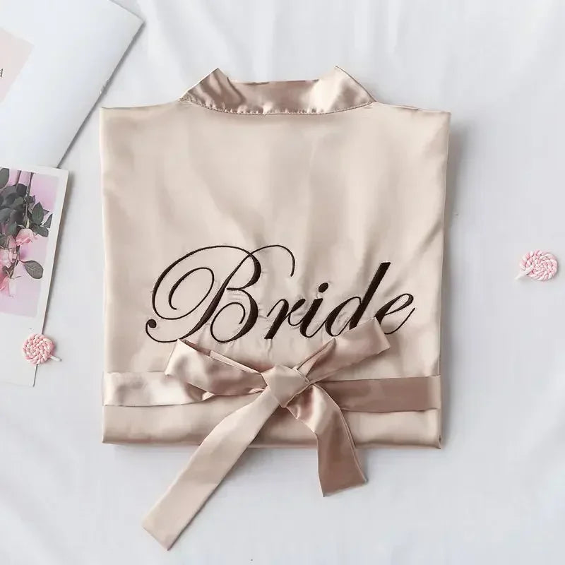 Folded satin robe with "Bride" in cursive, tied with a matching bow. Surrounding are floral cards and pink lollipops on a white surface.