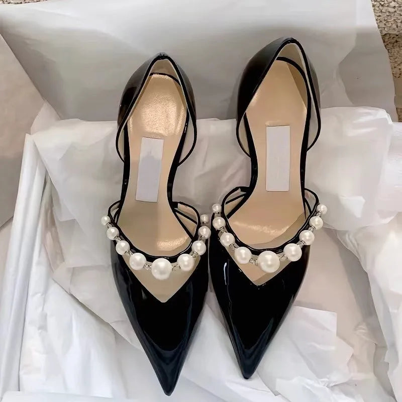 Elegant black patent leather high heels with pointed toes, adorned with pearl embellishments across the front. The shoes are displayed on white tissue paper inside a box.