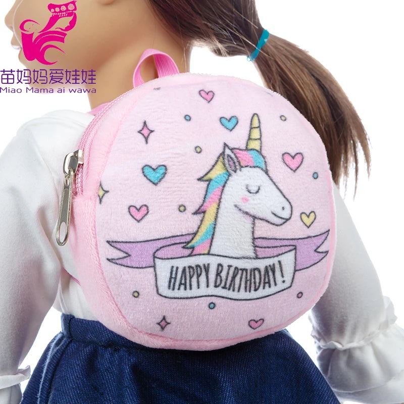 A doll wears a pink plush backpack with a colorful unicorn and "Happy Birthday!" text. The backpack features heart and star designs. The doll has a white shirt and blue skirt.