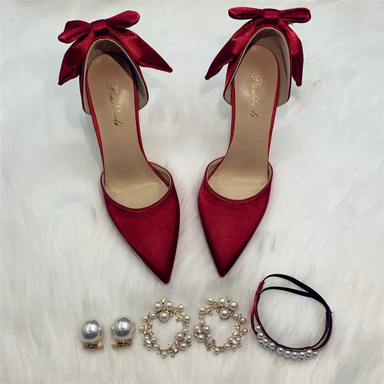 Red satin high heels with bows are placed on a white fur surface. Below them are pearl earrings, a pair of pearl hoops, and a pearl bracelet with a black ribbon.