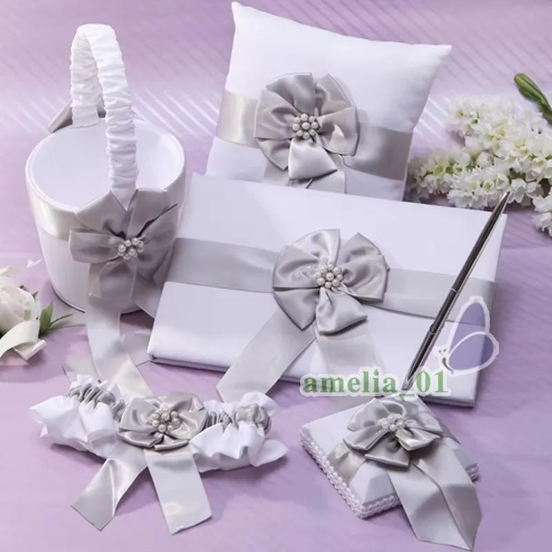 Elegant wedding accessory set with silver bows, including a white pillow, basket, guestbook, garter, and pen holder, displayed on a lavender background with flowers.