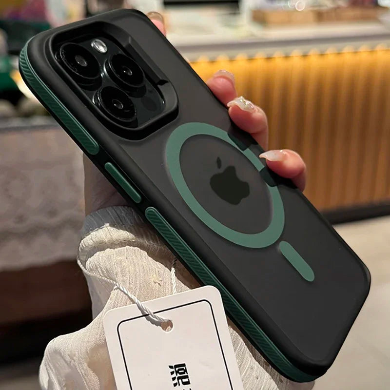 A hand holding a black iPhone with a green-bordered camera and MagSafe case. A tag is attached to the case. The background is softly blurred with warm lighting.