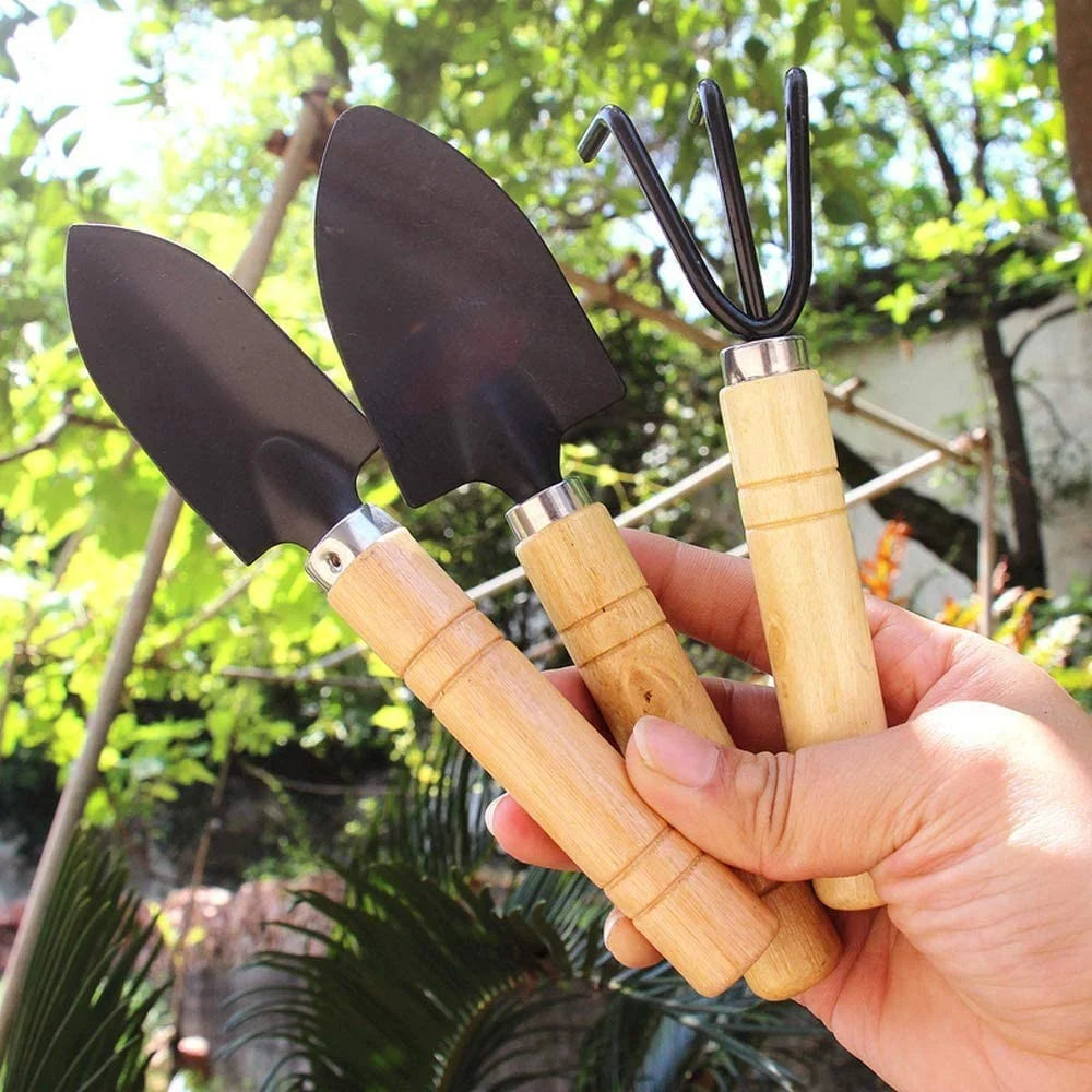 A hand holds three gardening tools with wooden handles: two small shovels and a three-pronged fork, set against a backdrop of green foliage in a sunny garden.