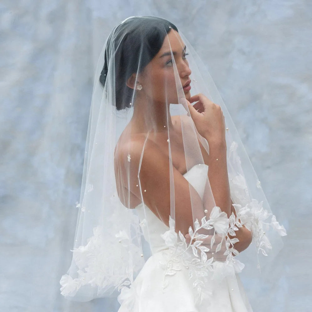 Bride in a white strapless gown and lace veil gazes forward thoughtfully against a soft, light blue background. Her hair is elegantly styled, and she wears subtle pearl earrings.