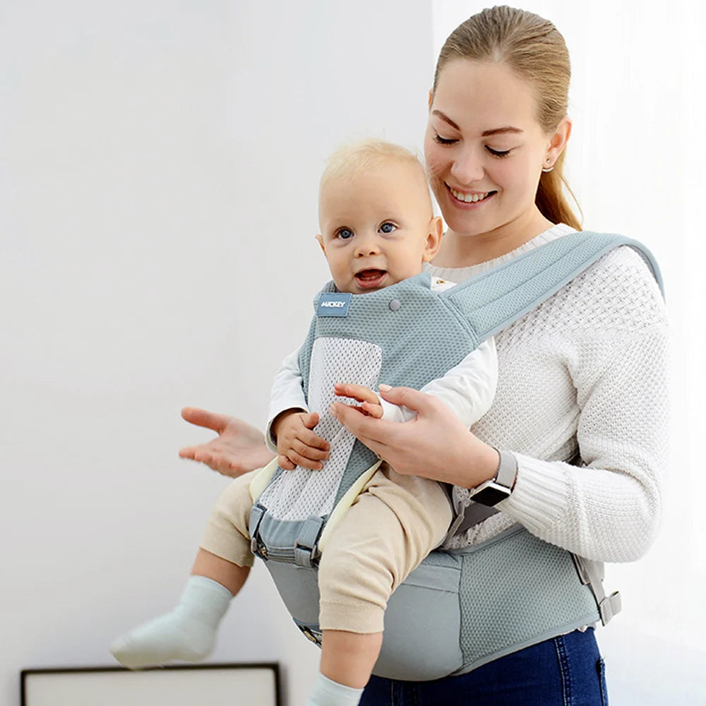 A smiling woman in a white sweater carries a baby in a blue front-facing carrier. The baby is also smiling, and they are in a bright, airy room.