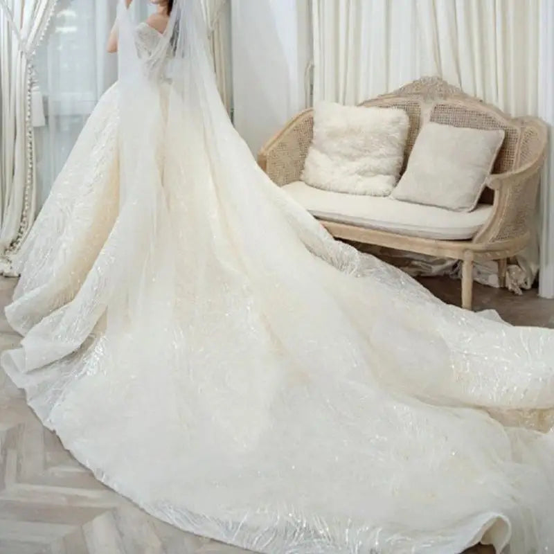 A bride in a flowing, embellished wedding gown with a long train stands near a wicker sofa with plush cushions, surrounded by light curtains and soft, natural light.